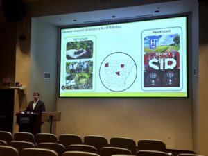 Dr. Anthony Gunderman presents during a UAMS Research & Innovation seminar, standing at a podium beside a large screen displaying research applications in agriculture and healthcare.