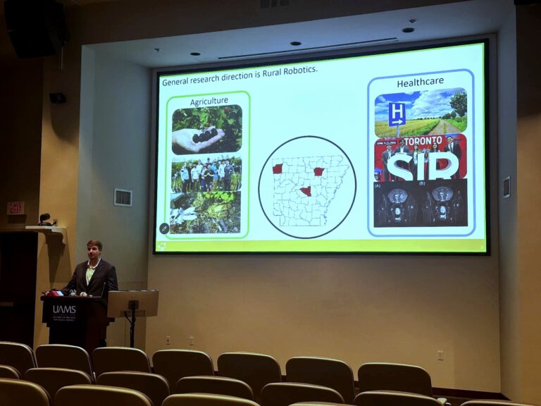 Dr. Anthony Gunderman presents during a UAMS Research & Innovation seminar, standing at a podium beside a large screen displaying research applications in agriculture and healthcare.